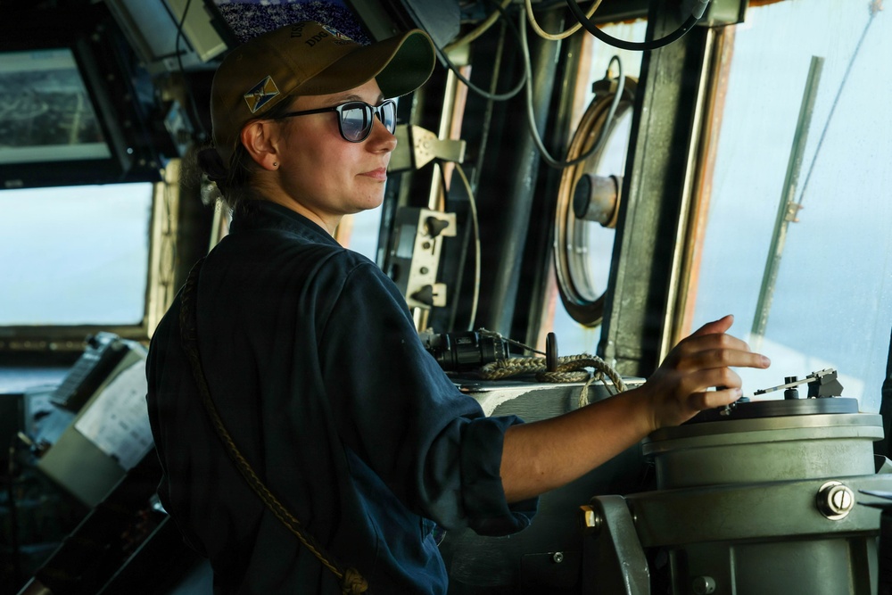 A female sailor or officer is stationed at the helm/bridge of a naval or military vessel, operating navigation equipment. She is wearing a dark blue uniform, sunglasses, and a tan/gold baseball cap with a star emblem. The setting is clearly a ship's bridge with open water visible through the windows.