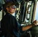 A female sailor or officer is stationed at the helm/bridge of a naval or military vessel, operating navigation equipment. She is wearing a dark blue uniform, sunglasses, and a tan/gold baseball cap with a star emblem. The setting is clearly a ship's bridge with open water visible through the windows.