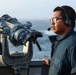 A naval personnel member stands on the deck of a ship at sea, wearing a communication headset and glasses while on watch duty. A large military-grade binocular/optical device marked 'US' is mounted on a railing in the foreground. The ocean is visible in the background under hazy sky conditions.