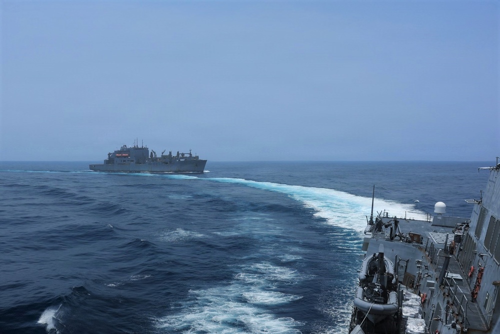 Two naval vessels are underway at sea, with a warship in the foreground on the right and a larger auxiliary/supply ship visible in the middle distance. Both vessels are moving through open ocean, leaving visible white wake patterns in the blue water. The image appears to depict a naval operation, possibly a replenishment-at-sea (RAS) approach or formation sailing.