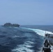 Two naval vessels are underway at sea, with a warship in the foreground on the right and a larger auxiliary/supply ship visible in the middle distance. Both vessels are moving through open ocean, leaving visible white wake patterns in the blue water. The image appears to depict a naval operation, possibly a replenishment-at-sea (RAS) approach or formation sailing.