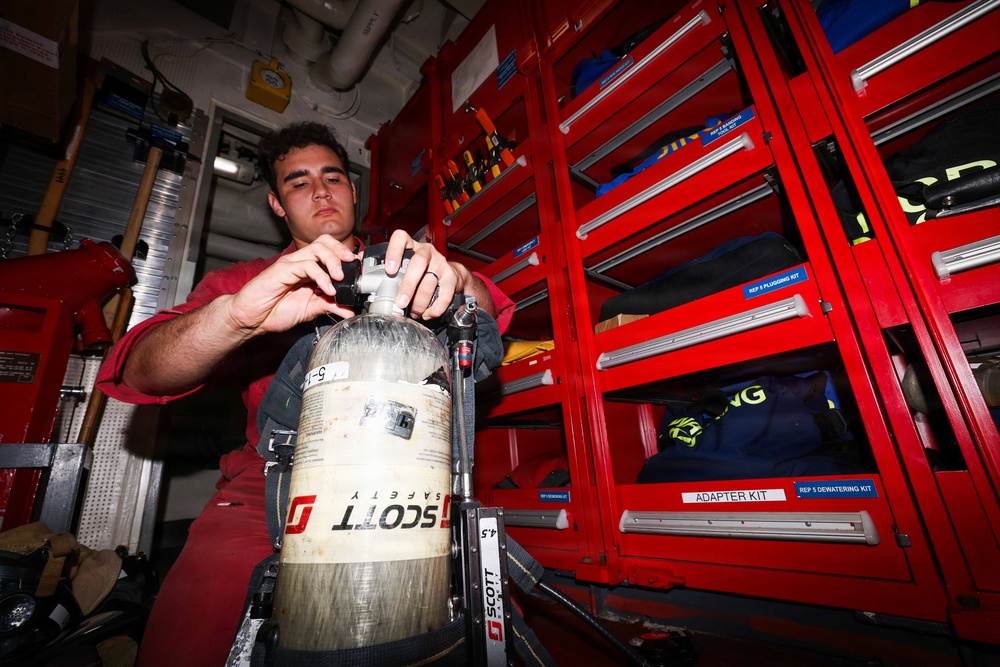 A young man in a red uniform is working with a Scott Safety SCBA (Self-Contained Breathing Apparatus) cylinder, appearing to inspect or connect the regulator valve. The setting appears to be inside a fire truck or emergency response vehicle, evidenced by the red tool storage cabinets with labeled drawers in the background. The environment is consistent with firefighting or emergency services equipment maintenance.