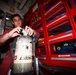 A young man in a red uniform is working with a Scott Safety SCBA (Self-Contained Breathing Apparatus) cylinder, appearing to inspect or connect the regulator valve. The setting appears to be inside a fire truck or emergency response vehicle, evidenced by the red tool storage cabinets with labeled drawers in the background. The environment is consistent with firefighting or emergency services equipment maintenance.
