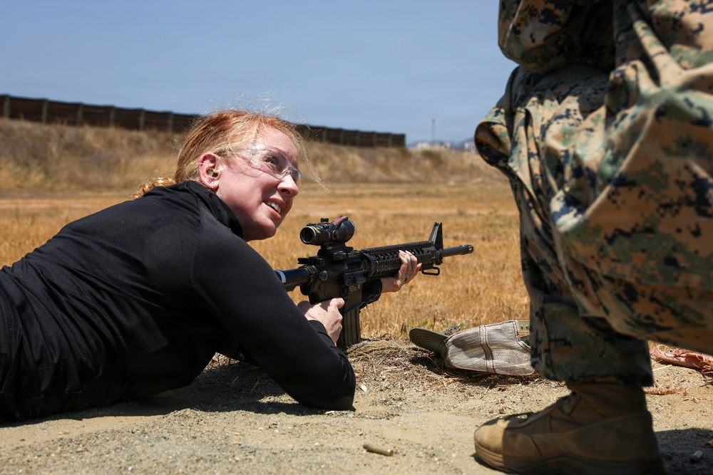 A woman in civilian clothing (black top) is in a prone shooting position on a firing range, aiming an M4-style carbine rifle equipped with an optical scope. She is wearing safety glasses and ear protection, suggesting a supervised training or demonstration exercise. A person in MARPAT (Marine Corps digital camouflage) uniform stands nearby, indicating a military training environment.