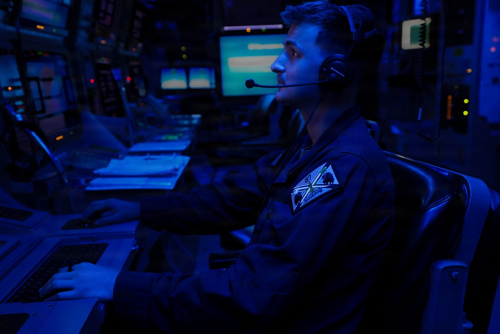 A military personnel member is seated at a multi-screen operations console in a darkened command or combat information center, illuminated primarily by blue ambient lighting. The individual is wearing a flight suit or coverall with a unit patch and a communications headset, actively monitoring screens and operating a keyboard. The environment is consistent with a naval combat information center (CIC) or similar military command station.