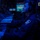 Routine Operations Aboard USS Mason (DDG 87) A military personnel member is seated at a multi-screen operations console in a darkened command or combat information center, illuminated primarily by blue ambient lighting. The individual is wearing a flight suit or coverall with a unit patch and a communications headset, actively monitoring screens and operating a keyboard. The environment is consistent with a naval combat information center (CIC) or similar military command station.