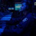 A military personnel member is seated at a multi-screen operations console in a darkened command or combat information center, illuminated primarily by blue ambient lighting. The individual is wearing a flight suit or coverall with a unit patch and a communications headset, actively monitoring screens and operating a keyboard. The environment is consistent with a naval combat information center (CIC) or similar military command station.