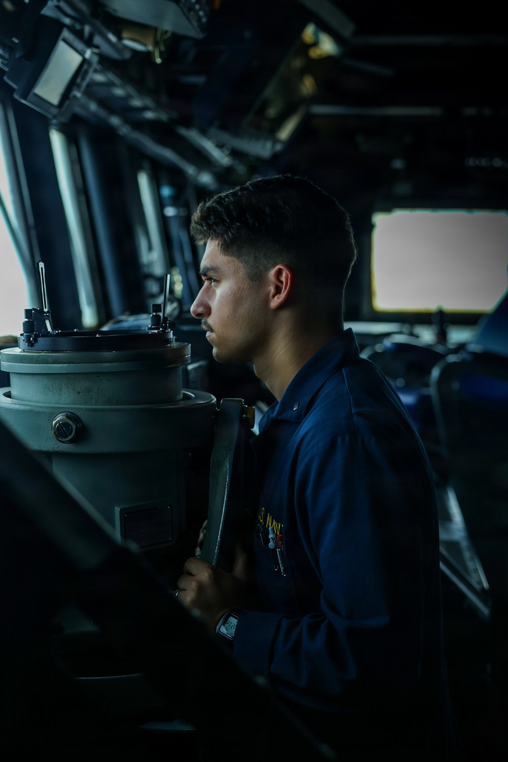 A young U.S. Navy sailor in a dark blue uniform is stationed at what appears to be a ship's bridge, looking through or operating a navigational instrument, likely a gyrocompass or bearing repeater. The setting is the interior of a naval vessel's pilothouse, filled with various electronic displays and control equipment. The image is dramatically lit with natural light coming through bridge windows.
