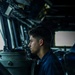 A young U.S. Navy sailor in a dark blue uniform is stationed at what appears to be a ship's bridge, looking through or operating a navigational instrument, likely a gyrocompass or bearing repeater. The setting is the interior of a naval vessel's pilothouse, filled with various electronic displays and control equipment. The image is dramatically lit with natural light coming through bridge windows.