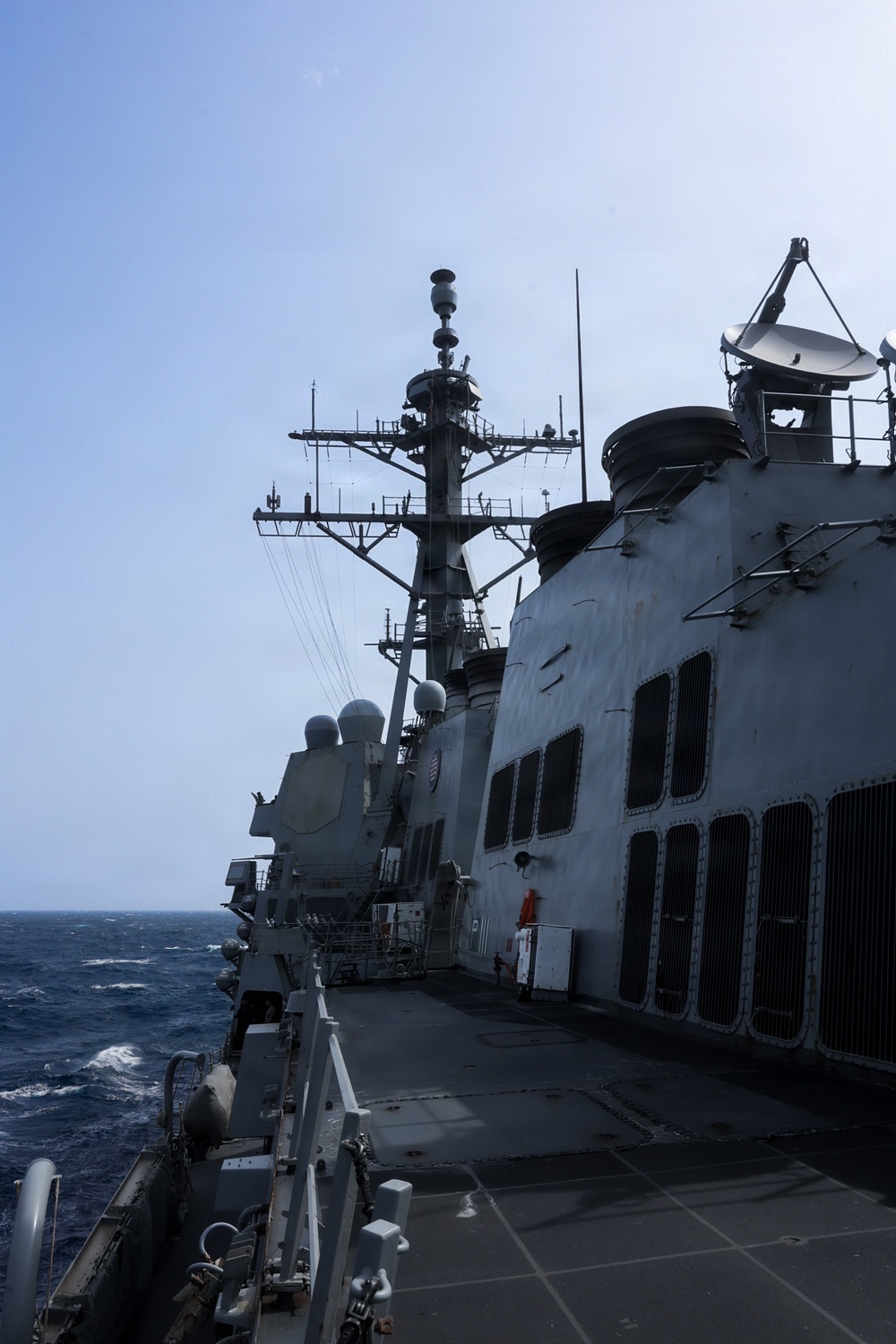 The image shows the superstructure and main mast of a U.S. Navy warship, likely an Arleigh Burke-class guided-missile destroyer, photographed from the main deck level. The vessel is underway at sea with moderate wave activity visible to port side. The photo captures the ship's communications and sensor equipment in detail against a hazy blue sky.