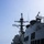 Routine Operations Aboard USS Mason (DDG 87) The image shows the superstructure and main mast of a U.S. Navy warship, likely an Arleigh Burke-class guided-missile destroyer, photographed from the main deck level. The vessel is underway at sea with moderate wave activity visible to port side. The photo captures the ship's communications and sensor equipment in detail against a hazy blue sky.