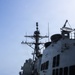The image shows the superstructure and main mast of a U.S. Navy warship, likely an Arleigh Burke-class guided-missile destroyer, photographed from the main deck level. The vessel is underway at sea with moderate wave activity visible to port side. The photo captures the ship's communications and sensor equipment in detail against a hazy blue sky.
