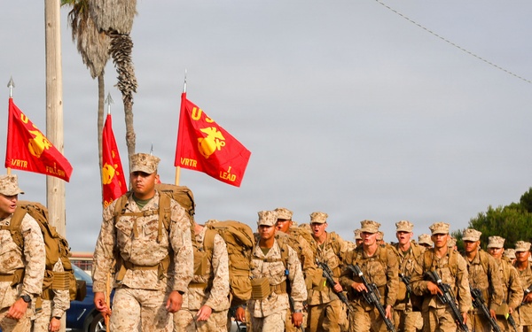 A column of U.S. Marines in desert MARPAT camouflage uniforms marches in formation along a paved road. The formation is led by officers or senior NCOs carrying red U.S. Marine Corps guidon flags. The setting appears to be a military base or coastal installation, with palm trees and civilian vehicles visible in the background.
