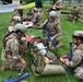 Military personnel in camouflage uniforms and helmets with green markings are conducting a combat casualty care training exercise on a grassy field. Soldiers are practicing tactical field care on simulated casualties using litters, moulage (simulated wounds), and medical equipment. The scene includes multiple training stations with instructors and observers present in the background.