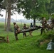 A group of military personnel in camouflage uniforms and helmets are carrying what appears to be a litter or stretcher with a casualty or simulated casualty across a grassy area. The scene appears to be a training exercise or field operation near a fenced perimeter. Several individuals in mixed attire, including civilian-style clothing, are assisting with the carry.