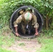 A soldier in full combat gear is crawling through a large black corrugated drainage tunnel, emerging from the exit end. The setting appears to be a military obstacle course or training exercise conducted in a vegetated outdoor environment. A green silhouette target is visible to the left of the tunnel.