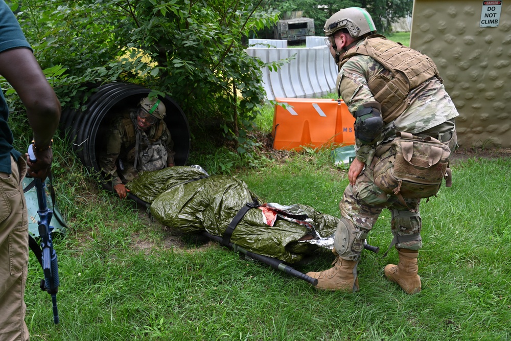 Military personnel in full combat gear are conducting a casualty evacuation training exercise. One soldier is emerging from a large drainage culvert pipe while pulling a litter (stretcher) carrying a simulated casualty wrapped in a thermal/emergency blanket with visible simulated blood. A second soldier in OCP (Operational Camouflage Pattern) uniform assists from outside the pipe.