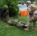 Military personnel in full combat gear are conducting a casualty evacuation training exercise. One soldier is emerging from a large drainage culvert pipe while pulling a litter (stretcher) carrying a simulated casualty wrapped in a thermal/emergency blanket with visible simulated blood. A second soldier in OCP (Operational Camouflage Pattern) uniform assists from outside the pipe.