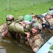 Military personnel in full combat gear are conducting a water obstacle training exercise, carrying what appears to be a simulated casualty through a shallow water channel. Multiple soldiers in camouflage uniforms and helmets are working together to transport the 'casualty' wrapped in a poncho or tarp. One soldier in the background maintains a security posture with a training weapon while others focus on the casualty extraction.