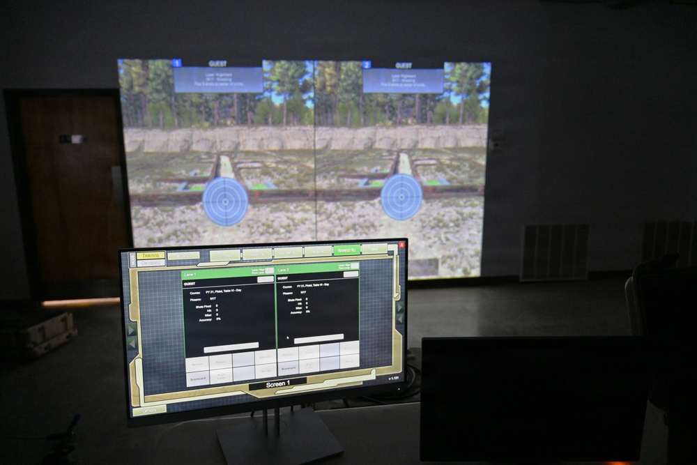 The image shows a firearms simulation training facility with a large projected screen displaying a two-lane virtual shooting range with circular targets in an outdoor woodland environment. A control monitor labeled 'Screen 1' in the foreground displays a management interface showing Lane 1 and Lane 2 with player statistics and scoring data. The setup appears to be an indoor marksmanship simulator used for training purposes.