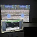 The image shows a firearms simulation training facility with a large projected screen displaying a two-lane virtual shooting range with circular targets in an outdoor woodland environment. A control monitor labeled 'Screen 1' in the foreground displays a management interface showing Lane 1 and Lane 2 with player statistics and scoring data. The setup appears to be an indoor marksmanship simulator used for training purposes.