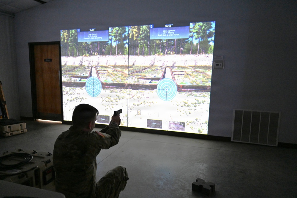 A soldier in military camouflage uniform is kneeling and aiming a pistol at a large projected simulation screen showing two virtual shooting range targets. The screen displays a split-screen interface labeled 'GUEST 1' and 'GUEST 2' with circular bullseye targets in an outdoor range environment. This appears to be a military marksmanship training simulator, likely a Engagement Skills Trainer (EST) or similar system.