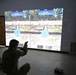 A soldier in military camouflage uniform is kneeling and aiming a pistol at a large projected simulation screen showing two virtual shooting range targets. The screen displays a split-screen interface labeled 'GUEST 1' and 'GUEST 2' with circular bullseye targets in an outdoor range environment. This appears to be a military marksmanship training simulator, likely a Engagement Skills Trainer (EST) or similar system.