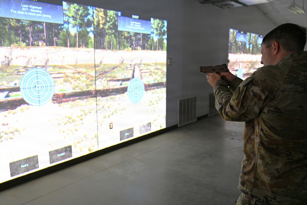 A military service member in MultiCam/OCP camouflage uniform is using a firearms simulation training system, aiming a pistol (appears to be an M17) at projected circular targets on a large screen. The simulation displays an outdoor woodland/field environment with bullseye targets and on-screen text indicating 'Laser Alignment' and 'M17 - Standing' mode. This appears to be a virtual marksmanship training facility used for weapons qualification and zeroing exercises.