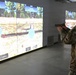 A military service member in MultiCam/OCP camouflage uniform is using a firearms simulation training system, aiming a pistol (appears to be an M17) at projected circular targets on a large screen. The simulation displays an outdoor woodland/field environment with bullseye targets and on-screen text indicating 'Laser Alignment' and 'M17 - Standing' mode. This appears to be a virtual marksmanship training facility used for weapons qualification and zeroing exercises.
