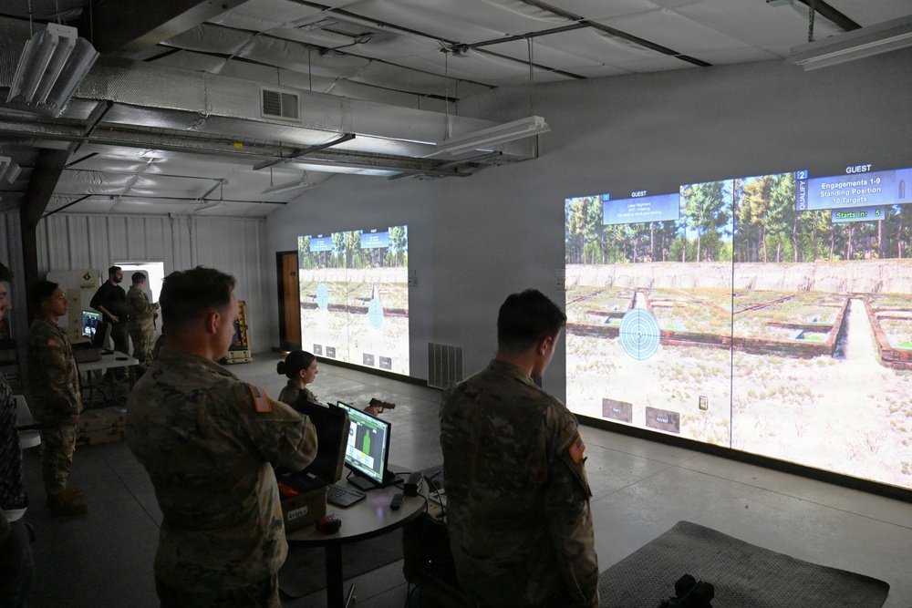Military personnel are gathered in an indoor virtual marksmanship training facility, observing and operating a large-screen shooting simulator. The room features multiple projected screens displaying a virtual outdoor firing range with targets. A seated operator at a workstation appears to be controlling or monitoring the simulation while soldiers observe.