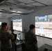 Military personnel are gathered in an indoor virtual marksmanship training facility, observing and operating a large-screen shooting simulator. The room features multiple projected screens displaying a virtual outdoor firing range with targets. A seated operator at a workstation appears to be controlling or monitoring the simulation while soldiers observe.
