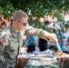 A U.S. Army soldier identified by name tape as 'RAUCH' is serving food at what appears to be a community outreach or volunteer event. The soldier, wearing OCP (Operational Camouflage Pattern) uniform with a U.S. flag patch, is using tongs to serve items from a food service setup to civilians wearing matching teal/turquoise shirts. The setting is outdoors under tree cover, suggesting a casual community engagement or charity event.