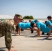 A smiling military servicemember in OCP (Operational Camouflage Pattern) uniform leads a group of young civilians in doing push-ups on an outdoor paved surface. The civilians are wearing matching turquoise/teal shirts and caps, suggesting they are part of an organized youth program or event. The setting appears to be a military installation, likely an airfield or tarmac area.
