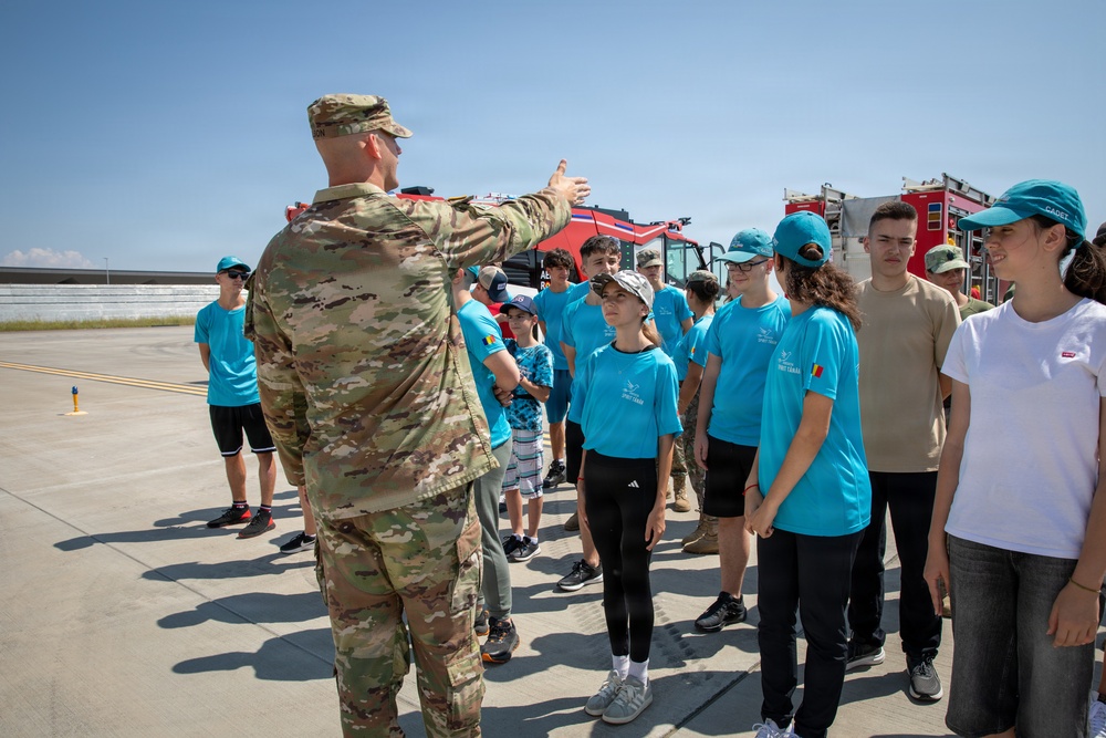 A military personnel in camouflage uniform is briefing or instructing a group of young people wearing matching teal/turquoise t-shirts on what appears to be an airfield tarmac. The youth group, likely cadets or students on a military base tour, are attentively listening while standing near a large red emergency/fire vehicle in the background. The setting suggests an organized educational visit or youth program at a military installation.