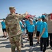 A military personnel in camouflage uniform is briefing or instructing a group of young people wearing matching teal/turquoise t-shirts on what appears to be an airfield tarmac. The youth group, likely cadets or students on a military base tour, are attentively listening while standing near a large red emergency/fire vehicle in the background. The setting suggests an organized educational visit or youth program at a military installation.