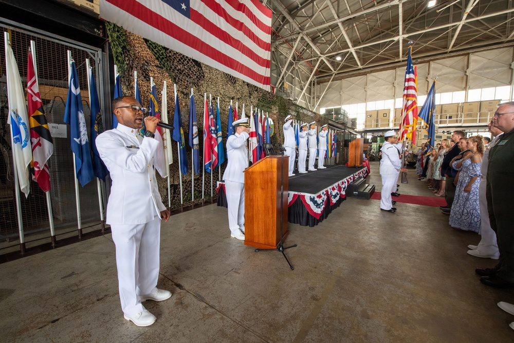 A formal U.S. Navy military ceremony is taking place inside a large hangar facility. A sailor in dress whites is performing vocals into a microphone in the foreground, while officers stand at attention on a decorated stage behind a wooden podium. A large American flag is prominently displayed on the back wall alongside numerous state and military flags.