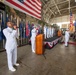 A formal U.S. Navy military ceremony is taking place inside a large hangar facility. A sailor in dress whites is performing vocals into a microphone in the foreground, while officers stand at attention on a decorated stage behind a wooden podium. A large American flag is prominently displayed on the back wall alongside numerous state and military flags.
