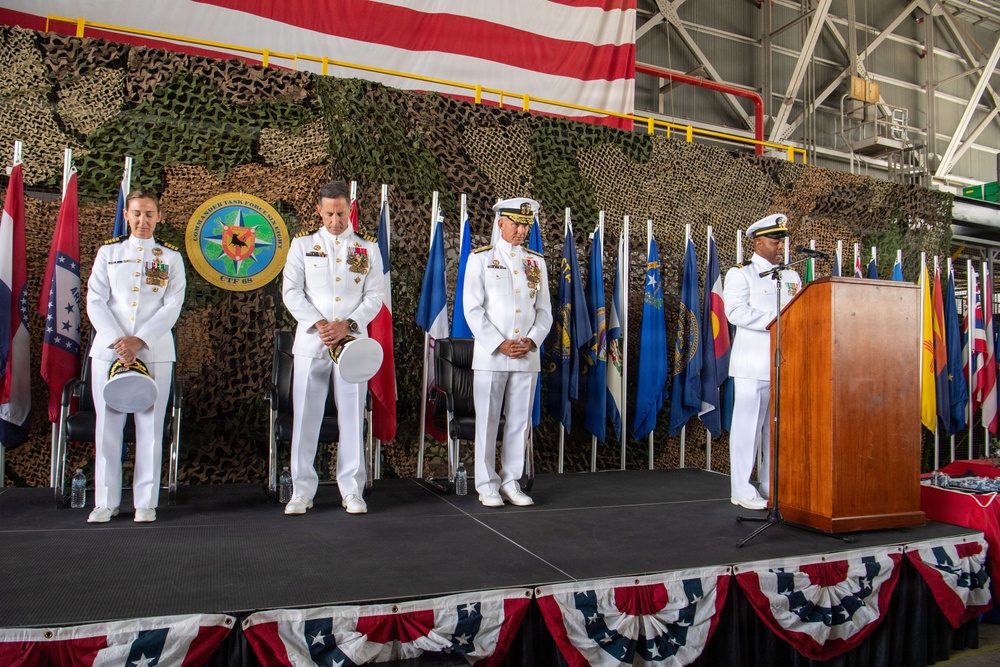 A formal U.S. Navy military ceremony, likely a change of command, taking place in a hangar facility. Four naval officers in dress white uniforms are on a raised stage, with three standing in a bowed/reverent posture and one at a podium, suggesting a prayer or invocation is being delivered. The stage is decorated with American flag bunting, state flags, a large U.S. flag backdrop, camouflage netting, and a Commander Task Force (CTF 68) seal.
