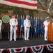A formal U.S. Navy military ceremony, likely a change of command, taking place in a hangar facility. Four naval officers in dress white uniforms are on a raised stage, with three standing in a bowed/reverent posture and one at a podium, suggesting a prayer or invocation is being delivered. The stage is decorated with American flag bunting, state flags, a large U.S. flag backdrop, camouflage netting, and a Commander Task Force (CTF 68) seal.