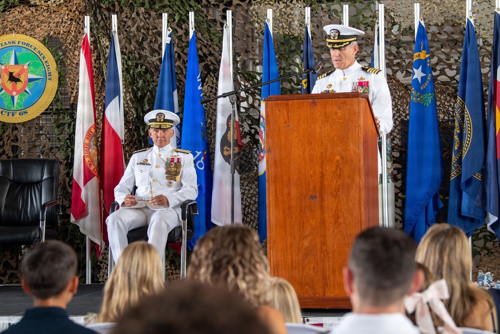 A formal U.S. Navy military ceremony, likely a change of command, is taking place on a decorated stage. Two senior naval officers in white dress uniforms are present — one speaking at a wooden podium and one seated — with an audience visible in the foreground. The backdrop features camouflage netting, multiple state and military flags, and a CTF 68 (Combined Task Force 68) emblem.
