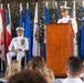 A formal U.S. Navy military ceremony, likely a change of command, is taking place on a decorated stage. Two senior naval officers in white dress uniforms are present — one speaking at a wooden podium and one seated — with an audience visible in the foreground. The backdrop features camouflage netting, multiple state and military flags, and a CTF 68 (Combined Task Force 68) emblem.