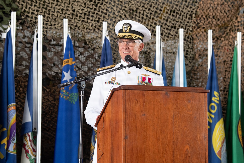 A senior U.S. Navy officer in dress white uniform is speaking at a podium during what appears to be a formal military ceremony. Multiple U.S. state flags are displayed behind him against a camouflage netting backdrop. The officer is wearing high-ranking insignia, medals, and a white combination cover consistent with flag officer rank.