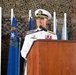 A senior U.S. Navy officer in dress white uniform is speaking at a podium during what appears to be a formal military ceremony. Multiple U.S. state flags are displayed behind him against a camouflage netting backdrop. The officer is wearing high-ranking insignia, medals, and a white combination cover consistent with flag officer rank.