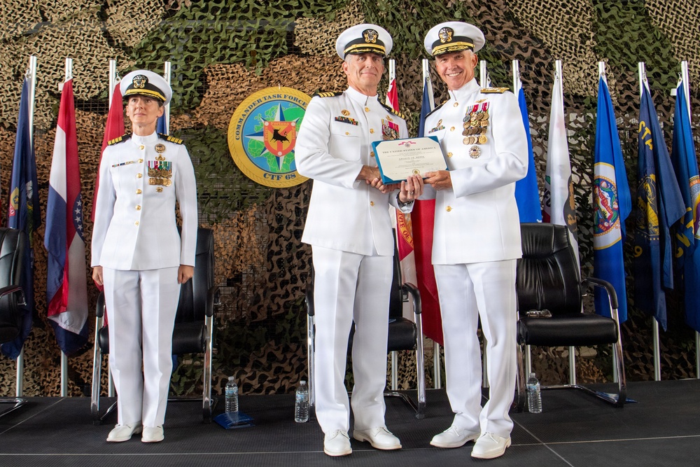 Three U.S. Navy officers in white dress uniforms are participating in a formal military ceremony, likely a change of command or award presentation. Two male officers in the center are jointly holding what appears to be a certificate or award document, while a female officer stands at attention to the left. The backdrop features a Commander Task Force 68 (CTF 68) emblem, camouflage netting, and multiple flags.