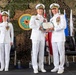 Three U.S. Navy officers in white dress uniforms are participating in a formal military ceremony, likely a change of command or award presentation. Two male officers in the center are jointly holding what appears to be a certificate or award document, while a female officer stands at attention to the left. The backdrop features a Commander Task Force 68 (CTF 68) emblem, camouflage netting, and multiple flags.