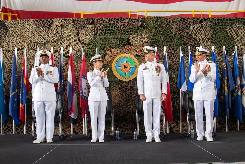 A formal military ceremony featuring four naval officers in white dress uniforms standing on a stage. A seal/emblem reading 'Commander Task Force' and 'CTF 65' is prominently displayed on the backdrop, surrounded by camouflage netting and multiple state/national flags. Three of the four officers are applauding, suggesting a change of command or award ceremony.