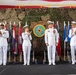 A formal military ceremony featuring four naval officers in white dress uniforms standing on a stage. A seal/emblem reading 'Commander Task Force' and 'CTF 65' is prominently displayed on the backdrop, surrounded by camouflage netting and multiple state/national flags. Three of the four officers are applauding, suggesting a change of command or award ceremony.