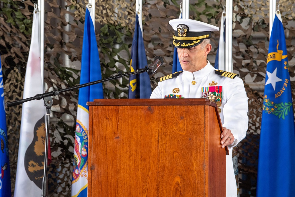 A senior U.S. Navy officer in white dress uniform is delivering a speech at a formal military ceremony from a wooden podium. Multiple state flags are visible in the background, including those of California and Nevada, along with camouflage netting as a backdrop. The officer displays numerous service ribbons and medals on his uniform, indicating high rank and extensive service.