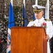 A senior U.S. Navy officer in white dress uniform is delivering a speech at a formal military ceremony from a wooden podium. Multiple state flags are visible in the background, including those of California and Nevada, along with camouflage netting as a backdrop. The officer displays numerous service ribbons and medals on his uniform, indicating high rank and extensive service.