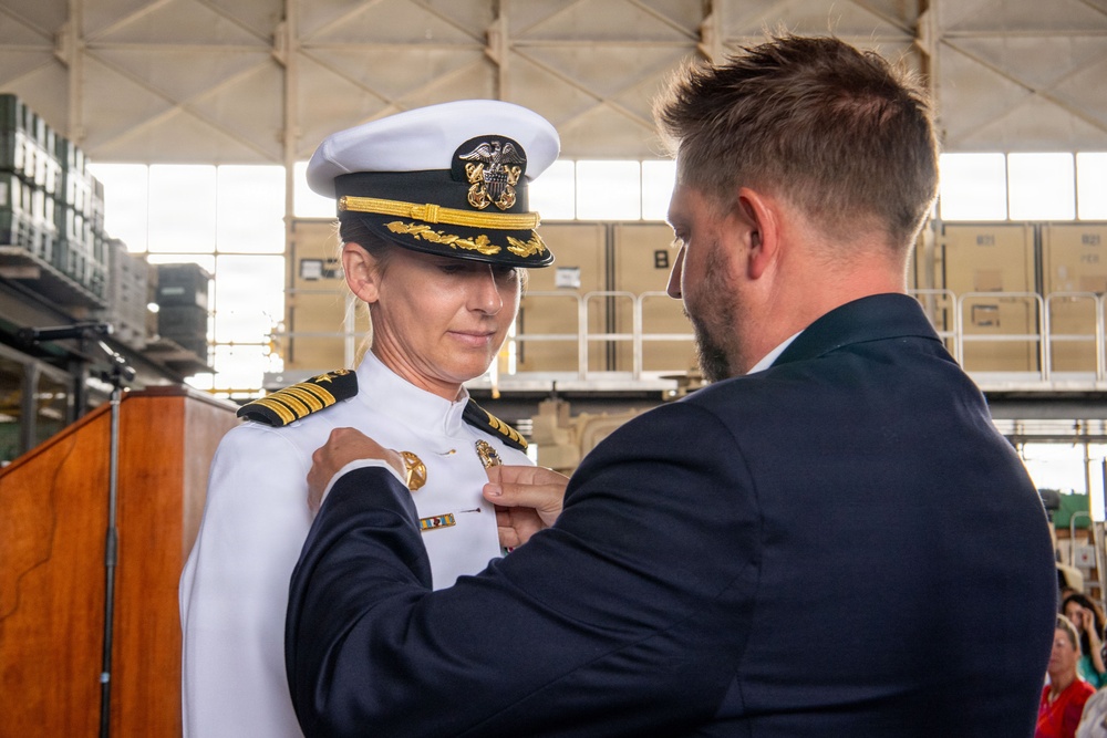 A military promotion or award ceremony is taking place, in which a civilian man is pinning a badge or insignia onto the uniform of a U.S. Navy officer. The officer is wearing a white dress uniform with gold shoulder boards indicating a senior rank (Captain or Commander), a white combination cover with gold scrambled eggs, and service ribbons. The setting appears to be a military hangar or pier facility.